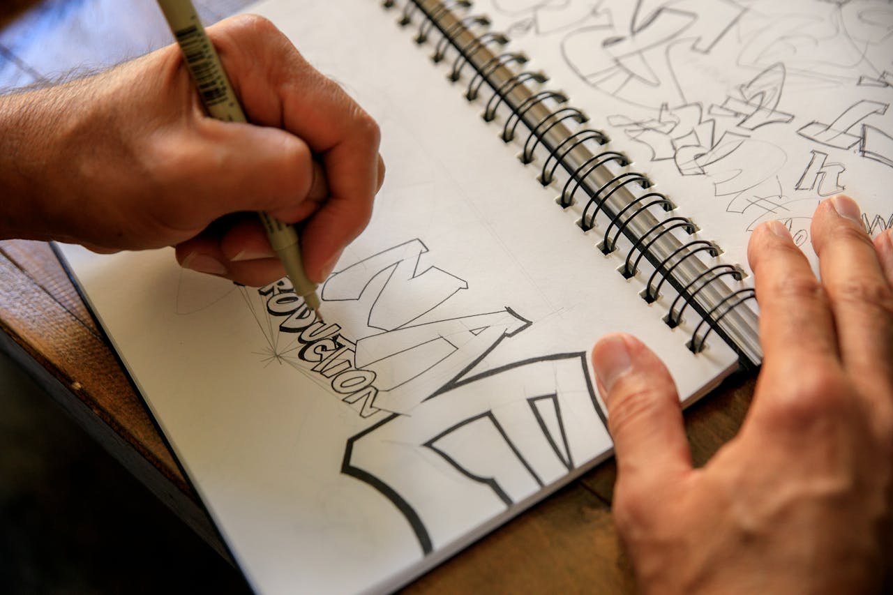 Close-up of hands sketching graffiti design in a spiral notebook on a table.